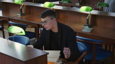 Kendini adamış bir öğrenci, kitaplar ve çalışma lambalarıyla çevrili sessiz bir kütüphane masasında okumaya dalmıştır. Medya