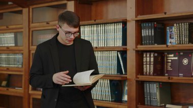 Düşünceli genç bir adam rahat ve davetkar bir kütüphanenin içinde raflarla çevrili bir kitap okumaya dalmış. Medya