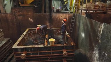 Dökümhane işçileri yoğun endüstriyel ortamda büyük bir küfe erimiş metal döküyorlar.