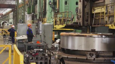 Fabrika işçisi, ağır bir sanayi ortamında makinenin üzerinde dönen büyük dairesel bir metal bileşeni izliyor.