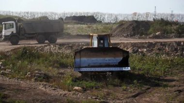 Sarı buldozer inşaat alanının tozunu atarken arka planda bir çöp kamyonu geçiyor. Kırp