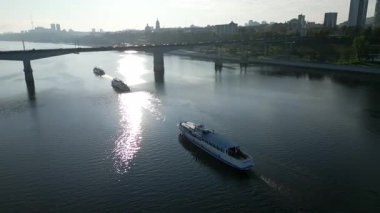 Beyaz yolcu feribotu gün doğumunda bir nehirde yol alırken, arka planda bir köprü ve şehir silüeti var. Kırp