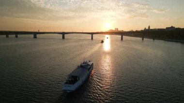 Beyaz yolcu feribotu gün batımında, bir köprü ve bir şehrin yakınında nehirden geçiyor. Kırp