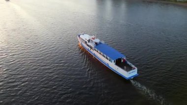 Beyaz ve mavi yolcu feribotu gün batımında, bir köprü ve şehir manzarası yakınında bir nehirde yol alıyor. Kırp