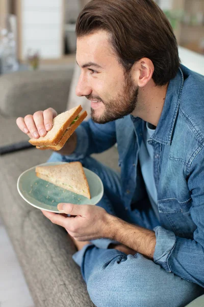 Χαρούμενος Εργένης Τρώει Σάντουιτς Πόδια Σταυρωμένα Στον Καναπέ — Φωτογραφία Αρχείου