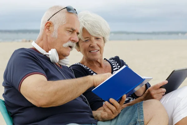 ビーチで本やタブレットを見ている老夫婦は — ストック写真