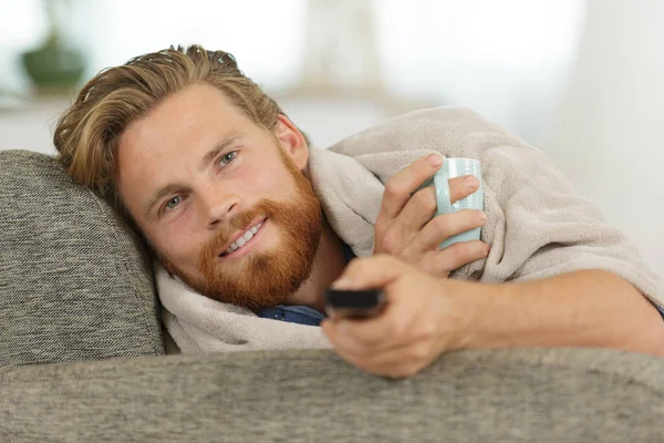 man under blanket on sofa with drink and remote control