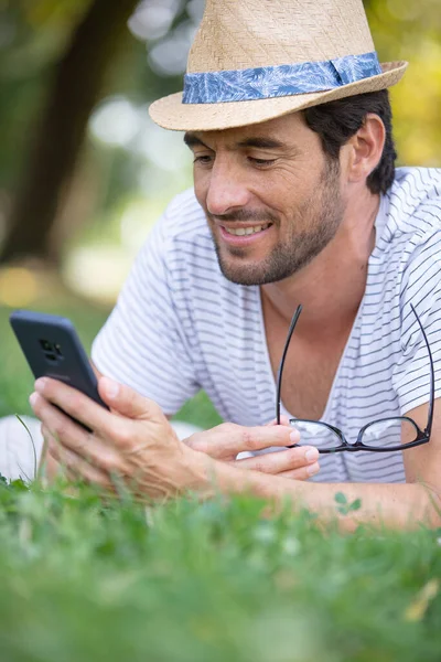 笑着的年轻人拿着手机躺在公园里 免版税图库照片