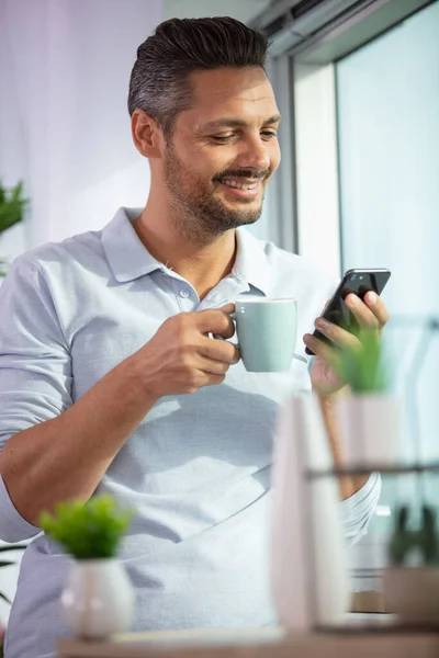 man stood by window holding cup looking at smartphone screen