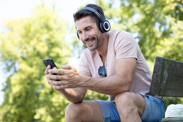 公园长椅上的中年男子 头戴耳机 手持智能手机 — 图库照片