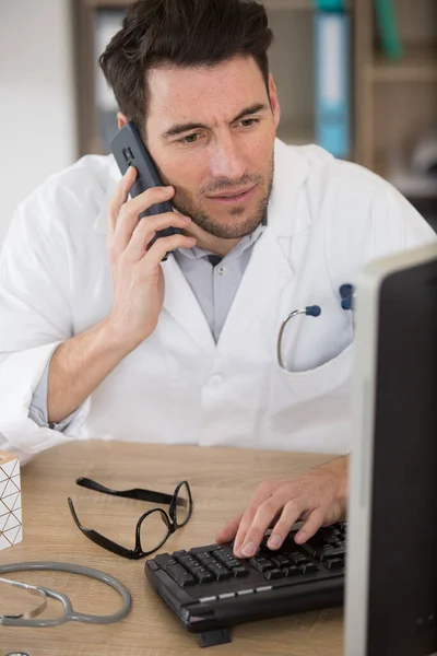 Άνδρας Γιατρός Που Χρησιμοποιεί Κινητό Τηλέφωνο Στην Κλινική Έναντι Υπολογιστή — Φωτογραφία Αρχείου