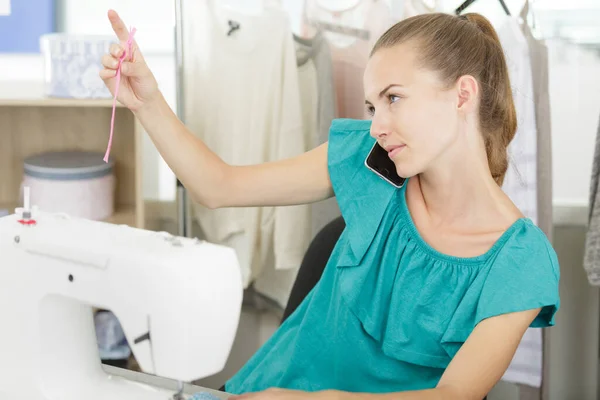 年轻女子在使用缝纫机的时候用智能手机说话 — 图库照片