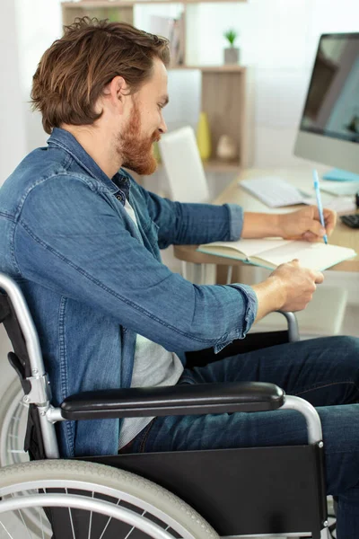 Άνθρωπος Αναπηρική Καρέκλα Γράφει Ένα Βιβλίο Στο Γραφείο Στο Σπίτι — Φωτογραφία Αρχείου