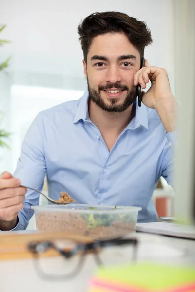 Przypadkowy Szczęśliwy Młody Człowiek Jedząc Sałatkę Podczas Rozmowy Telefonicznej — Zdjęcie stockowe