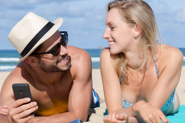 这对年轻夫妇用智能手机脸朝下躺在海滩上 — 图库照片