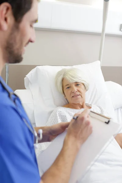 病院のベッドで高齢女性患者の近くに立つ男性医師は — ストック写真