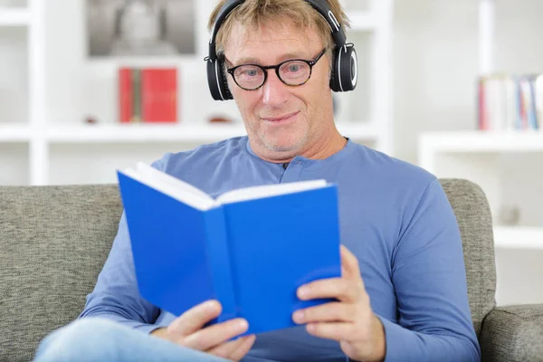ヘッドフォンをして家で読書をしている男は — ストック写真