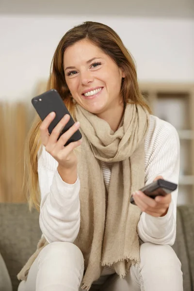 Cep Telefonuyla Gülümseyen Bir Kadın Televizyon Kumandasıyla Koltukta Oturuyor — Stok fotoğraf