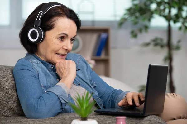在笔记本电脑上带着耳机聊天的老年妇女 — 图库照片