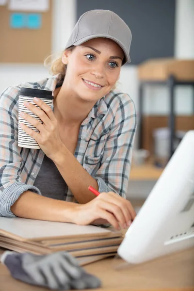 帽子をかぶった女性労働者がテイクアウトコーヒーを持ってコンピュータを使って — ストック写真