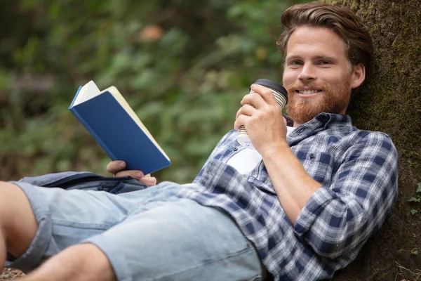 男人靠在树上看书 拿着外卖咖啡 — 图库照片