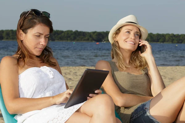 Δύο Γυναίκες Που Χρησιμοποιούν Τεχνολογία Ενώ Χαλαρώνουν Στην Παραλία — Φωτογραφία Αρχείου
