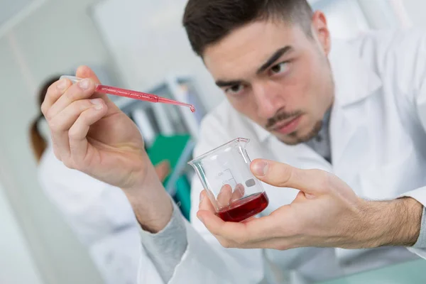 年轻的男性科学家将红色液体从吸盘滴入烧杯 — 图库照片