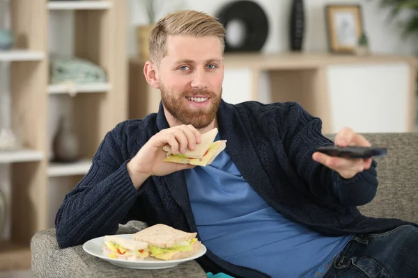坐在沙发上看电视和吃三明治的男人 — 图库照片