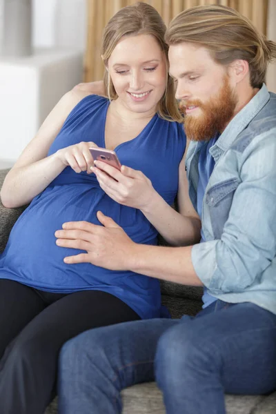年轻的怀孕夫妇用智能手机在沙发上放松 — 图库照片