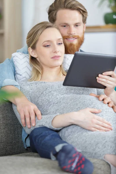 怀孕夫妇在沙发上放松地看着平板电脑 — 图库照片