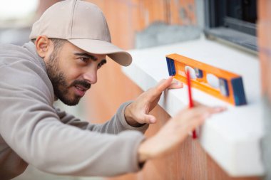 male builder using level tool to measure window outdoors clipart