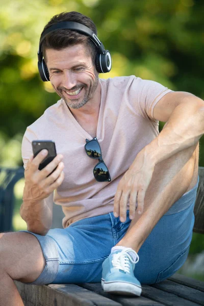 Hombre Riendo Sentó Banco Usando Auriculares Sosteniendo Teléfono Inteligente —  Fotos de Stock