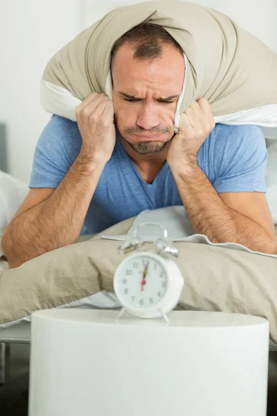 man holding pillow over ears to diminish alarm clock noise
