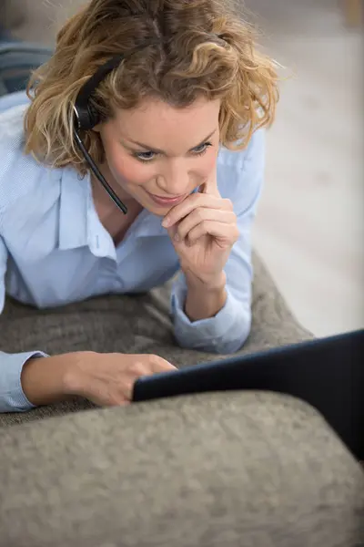 女人头戴耳机 用笔记本电脑躺在沙发上 免版税图库照片