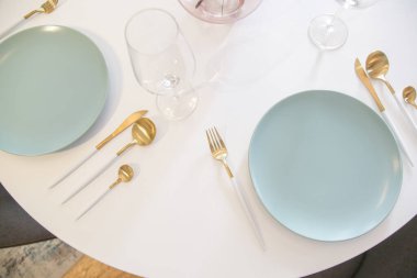 Dining table arrangement with ceramic blue plates, gold flatware set with white handles and wine glasses, top view. clipart