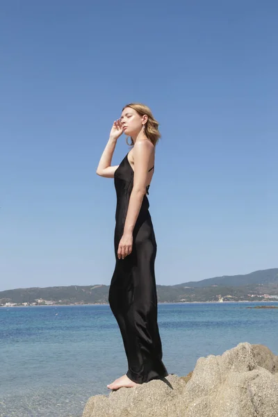 Outdoor portrait of female model in black silk dress on the seaside with beautiful calm sea behind, stylish and elegant summer fashion