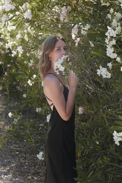 Outdoor portrait of female model in black silk dress near beautiful white flowering trees, stylish and elegant summer fashion