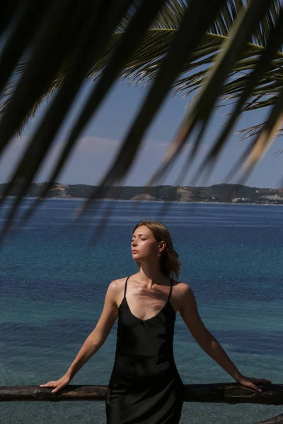 Outdoor portrait of female model in black silk dress on the seaside with beautiful calm sea behind, stylish and elegant summer fashion