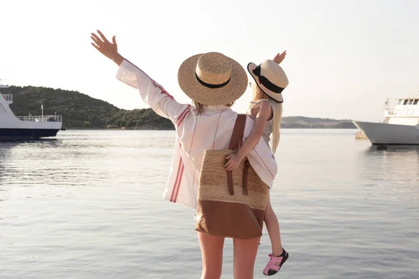 Mom and daughter with straw hats looking at the sea horizon, idyllic family summer vacation concept.
