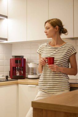 Young woman drinking coffee or tea from red mug in the kitchen