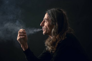 Studio portrait on black background of middle age man with long hair smoking cigarette