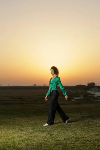 Fashion style portrait of young stunning woman posing in green silk blouse and denim trousers during sunset time