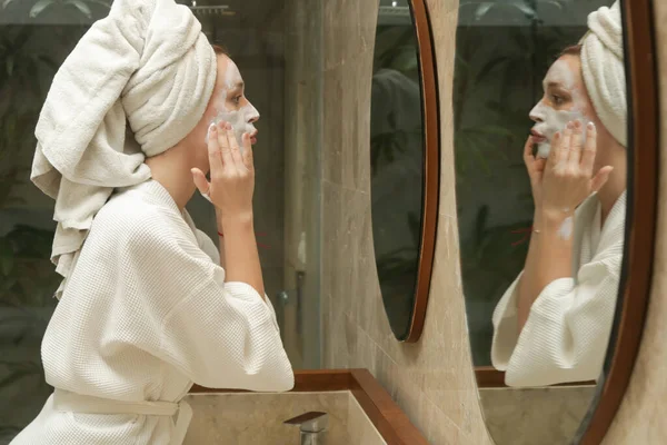 Woman applaying beauty face mask in front of the mirror, beauty skin care concept at home