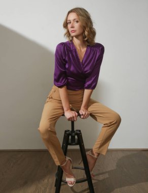 Stüdyo fotoğrafları, menekşe ipek saten bluz ve bej renkli yüksek paçalı pantolon giyen genç bir kadın model.