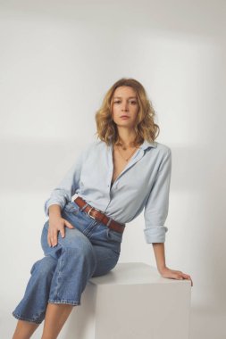 Beyaz arka planda açık mavi pamuklu gömlek ve mavi kot pantolon giyen genç bir kadının moda portresi.
