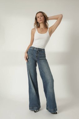 Beyaz atletli genç bir kadının moda portresi ve beyaz arka planda geniş bacaklı mavi kot pantolon.