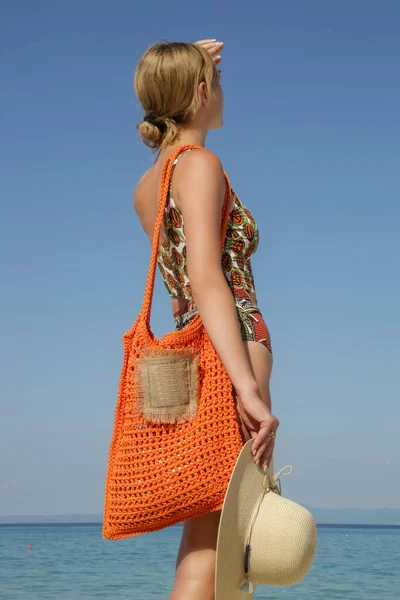 Young woman in tropical printed swimwear and straw hat cary orange crochet tote bag empty sandy beach on, summer fashion style, relaxed vacation