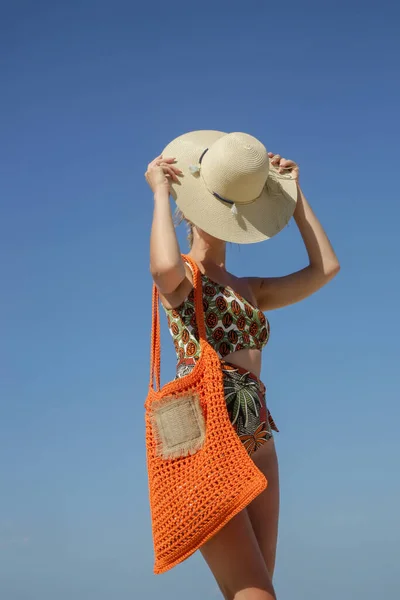 Young woman in tropical printed swimwear and straw hat cary orange crochet tote bag empty sandy beach on, summer fashion style, relaxed vacation