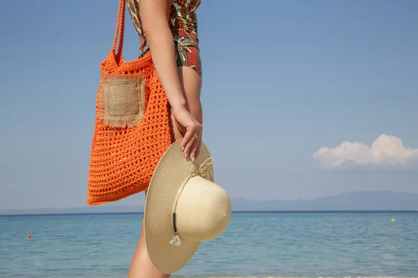 熱帯印刷された水着とわら帽子の若い女性ケアオレンジかぎ針編みトートバッグ空の砂浜オン 夏のファッションスタイル リラックスした休暇 ストック画像