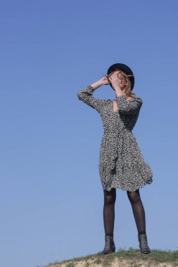 Boho Viscose çiçekli elbise ve arkasında açık mavi gökyüzü olan şapkalı kadın modelin serisi. Doğal ışıklandırmalı açık hava moda portresi.